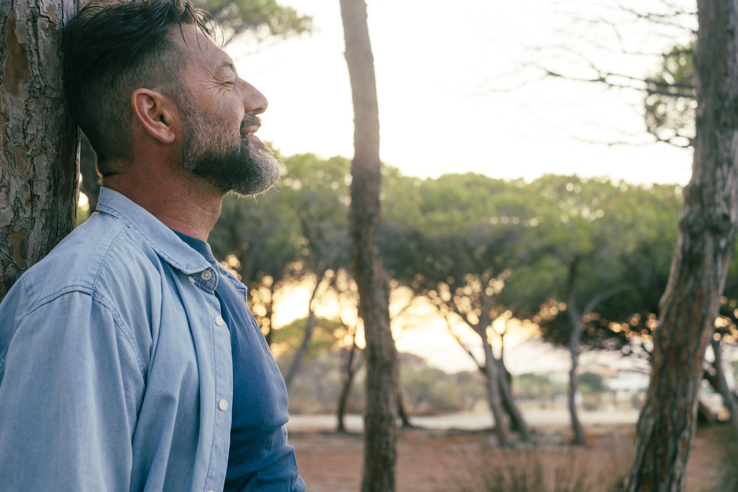 One adult man standing against a trunk tree having relaxation in outdoor leisure activity at the park alone. People and wellbeing connection with nature. Relax mental serene expression person mature