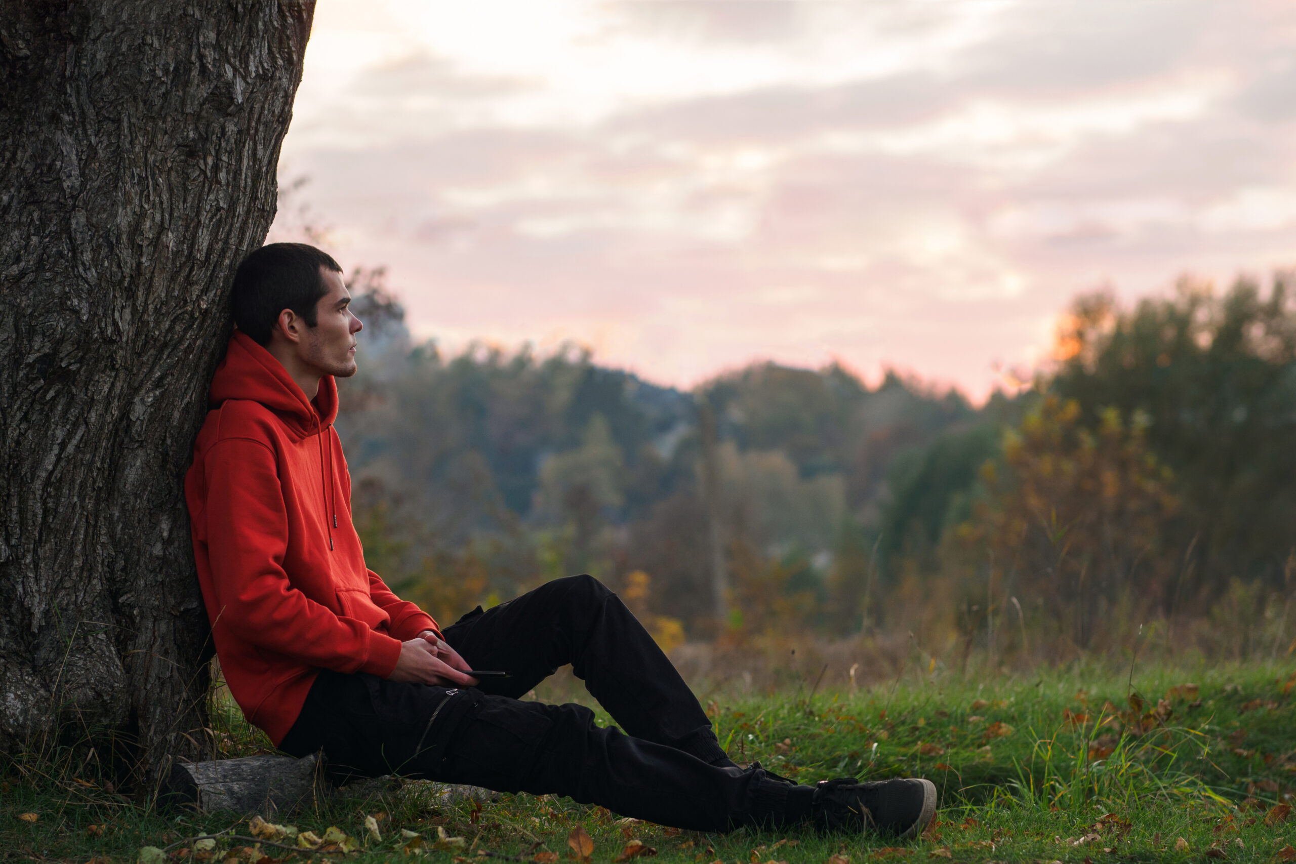A young man is sitting by a tree. Enjoys being alone . Autumn nature.